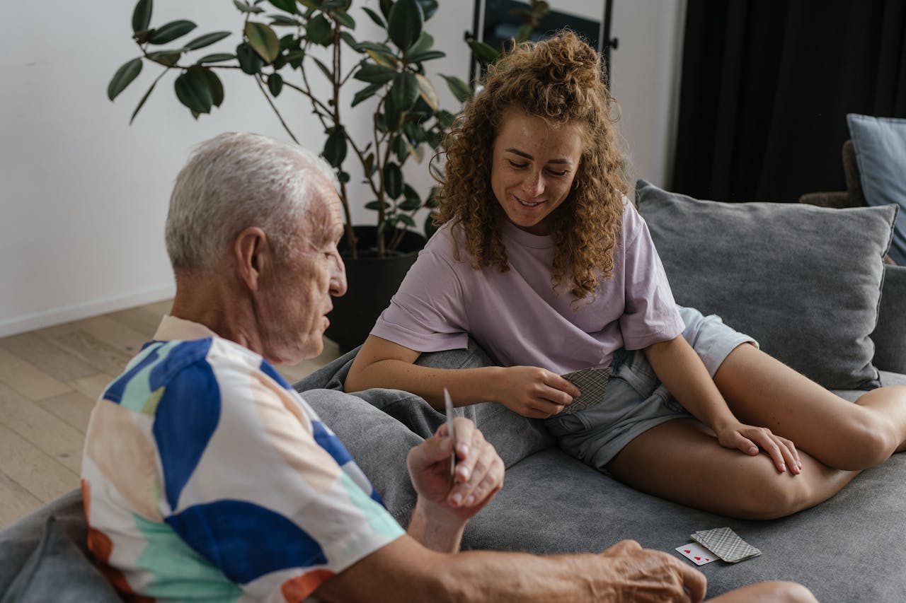 A senior adult and young woman share a joyful moment playing cards indoors on a cozy couch.